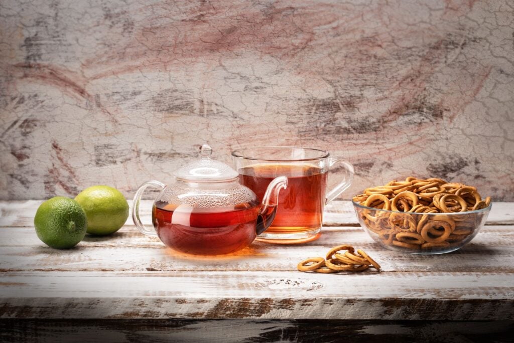 A glass teapot and cup with tea, accompanied by limes and snacks on a rustic wooden table.