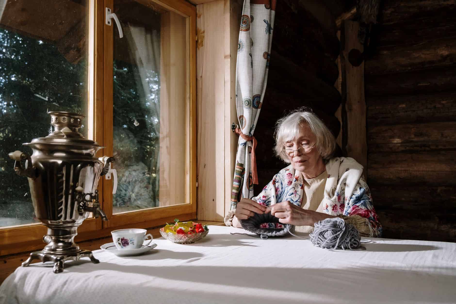 Elderly woman enjoys knitting in a rustic cabin with a samovar on a sunny day.
