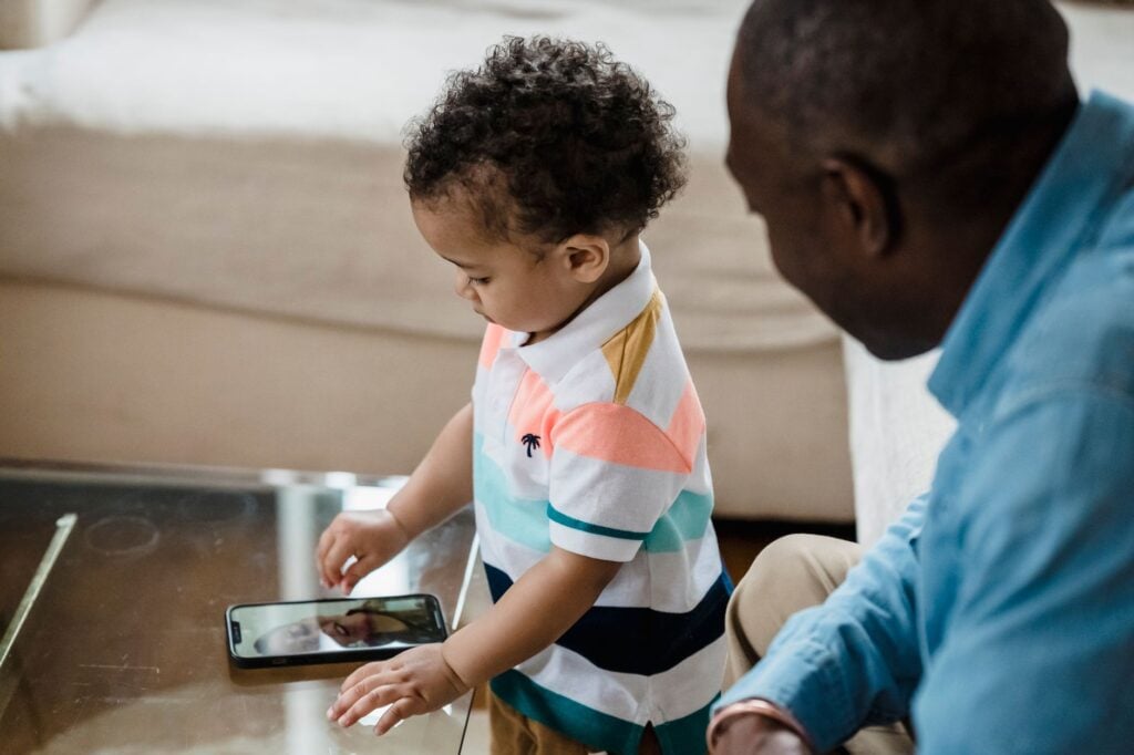 A father watches as his young son engages in a video call, reflecting modern family bonding indoors.