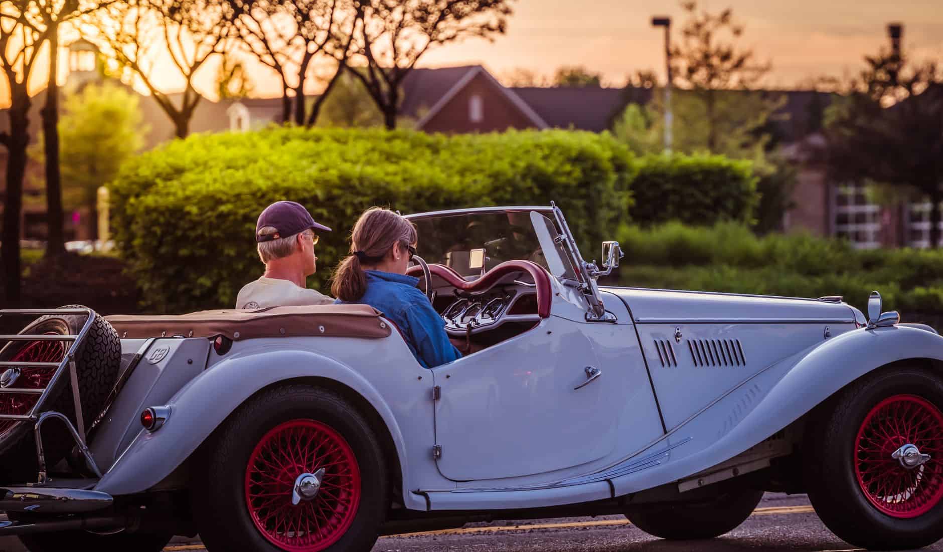 A couple drives a classic roadster on a scenic street at sunset, enjoying leisure and travel.