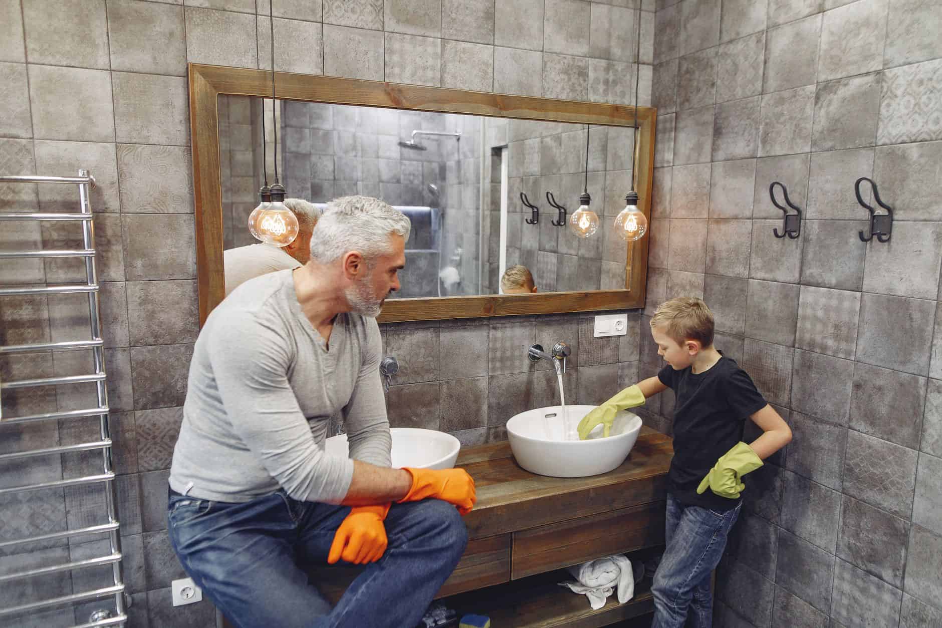 From above of grey haired dad with beard in orange rubber gloves teaching son tidying up bathroom